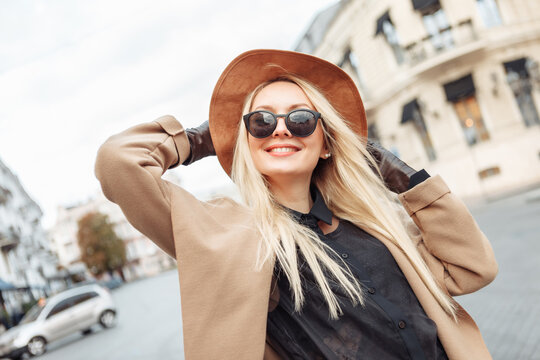 Portrait of young stylish blonde woman in autumn coat, sunglasses, gloves and felt hat in European city. Beautiful attractive girl