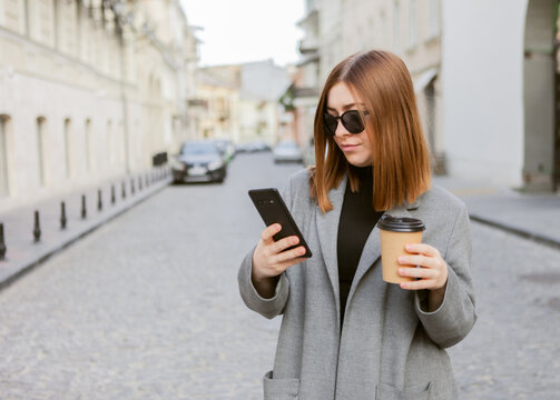 Young millennial woman with smooth hair dressed in an autumn coat and sunglasses uses a smartphone and holds a cup of coffee on the go in a European city.