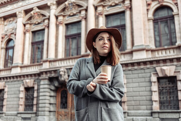 Fototapeta na wymiar Dramatic portrait of a fashionable woman in an autumn coat and hat. Attractive girl holding cup of coffee on the background of urban architecture