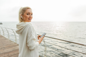 Fototapeta na wymiar Young fitness woman blonde in a white hoodie uses a smartphone and listens to music on headphones on the beach