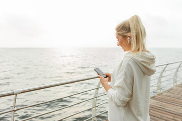 Naklejka premium Young fitness woman blonde in a white hoodie uses a smartphone and listens to music on headphones on the beach
