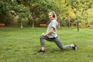 Fototapeta premium Young fit blonde woman warms up before training and listens to music on headphones in the park. Aerobica