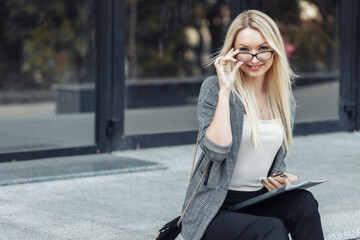 Fototapeta premium Business woman sitting on the steps during the break and holding folder and smartphone on the background of a business center