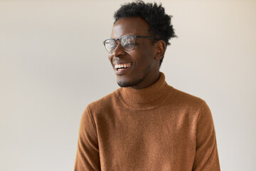 Obraz premium Attractive cheerful young Afro American man with stylish haircut expressing positive emotions, smiling broadly, being in good mood, posing isolated wearing eyeglasses and turtleneck sweater