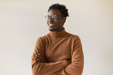 Studio shot of handsome enthusiastic dark skinned guy in trendy eyeglasses having inspired facial expression, thinking about future, making plans, keeping arms crossed on his chest and smiling