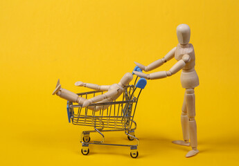 Shopping concept. Wooden puppets with supermarket trolley on yellow background. Spending time...
