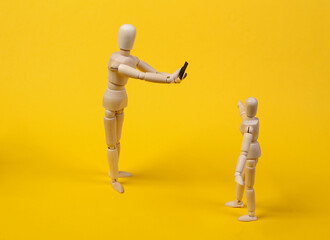 Two wooden puppets of a mannequin are photographed on a smartphone. Yellow studio background