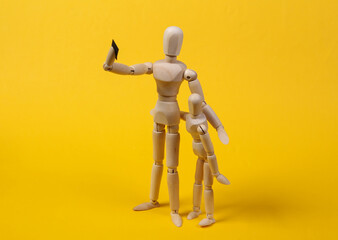 Two wooden puppets of a mannequin are photographed on a smartphone. Yellow studio background