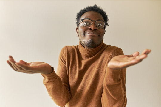 Studio image of emotional clueless young Afro American male in glasses, widening eyes and making helpless gesture with hands, having puzzled facial expression, can't make decision or choice