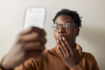 Isolated shot of funny emotional young black guy widening eyes and covering mouth with hand, expressing surprise while chatting online using front camera for video call, receiving shocking news