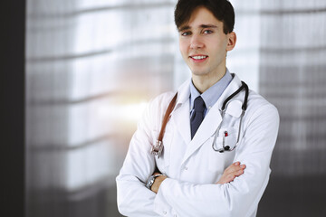 Cheerful smiling man-doctor standing with arms crossed in clinic. Perfect medical service with young smart physician in hospital. Medicine concept