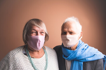 Dramatic light portrait of candid adorable happy senior couple of pensioners wearing medical face masks as protection against COVID-19 virus; concept of quarantine at home and outbreak prevention
