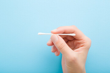 Young adult woman fingers holding stick of white cotton bud on light blue table background. Pastel color. Closeup. Top down view.