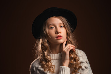 Charming blonde teen girl in black hat posing in studio on brown background