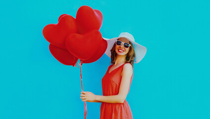 Portrait of beautiful happy smiling woman holding bunch of red heart shaped balloons wearing a summer straw hat and sunglasses on a blue background