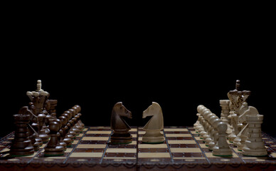 Chess faceoff of both knight horses on top of a chess board in front of a dark background surrounded by the king, queen, bishop, rook and pawns in the starting set up