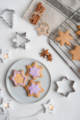 Decorating of sweet savory ginger cookies or gingerbread with violet icing on gray plate cooked for winter holidays on white wooden table with textile, cutters, cinnamon and grate. Vertical, flat lay