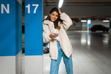 Beautiful girl in wireless earphones and mobile phone in hands in the underground parking. Portrait of modern young woman with smartphone in hands