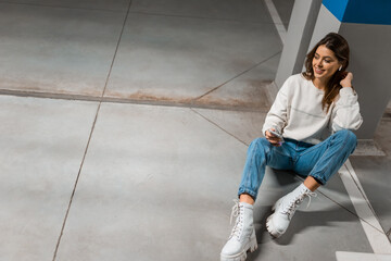 Stylish girl in the underground parking sits on the floor and listens to music on wireless headphones from smartphone