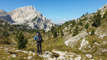 Fototapeta na wymiar A man with a big hiking backpack hiking in Italian Dolomites. He walks on a very narrow pathway. There are high and stony mountain chains in front. The slopes are overgrown with a few trees. Solitude