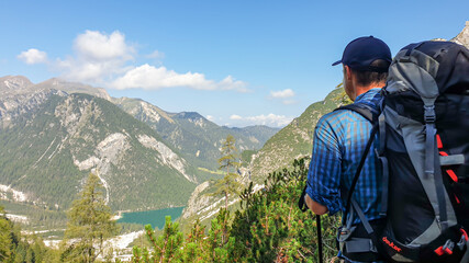 Fototapeta na wymiar A man with a big hiking backpack enjoying the distant view on the Pragser Wildsee, a lake in South Tyrolean Dolomites. There are high mountain chains around the lake. Serenity and calmness.