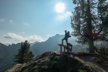 A man with hiking backpack standing on the wooden bench and spreading his arms wide. He enjoys the panoramic view on Italian Dolomites. Bright and sunny day. Few trees growing around it. Freedom