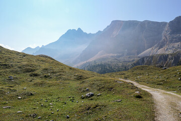 Fototapeta premium A gravelled road leading to vast valley in Italian Dolomites. High and sharp mountains in the back, with distinctive red color. Meadow with many boulders in front. Remote and desolate place. Serenity
