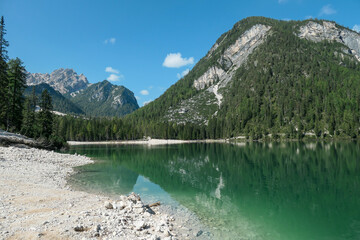 Naklejka na ściany i meble A panoramic view on the Pragser Wildsee, a lake in South Tyrolean Dolomites. High mountain chains around the lake. The sky and mountains are reflecting in the lake. Dense forest at the shore. Serenity