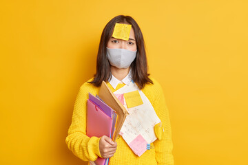 Upset female student works distantly during quarantine wears protective mask adhesive note with drawn graphic prepares project work looks sadly at camera isolated over yellow background. Coronavirus