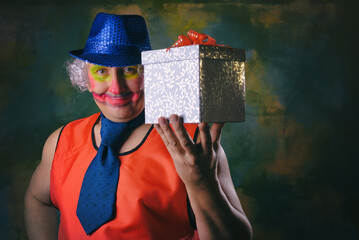 The clown holds a gift box in his hands. Grimaces and grimaces. Painted face.