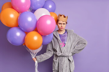 Fototapeta na wymiar Horizontal shot of unhappy woman dressed in soft dressing gown and paper crown holds bunch of colorful balloons frowns face isolated over purple background. Frustrated female has spoiled party
