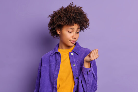 Unhappy young Afro American woman looks at her nails wants to make new manicure dressed in stylish clothing isolated over vivid purple background. Monochrome shot. Female concentrated at hand