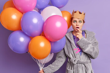 Fototapeta na wymiar Impressed female model stares bugged eyes at camera feels surprised holds colorful inflated balloons dressed in domestic gown going to celebrate something isolated over purple studio background.