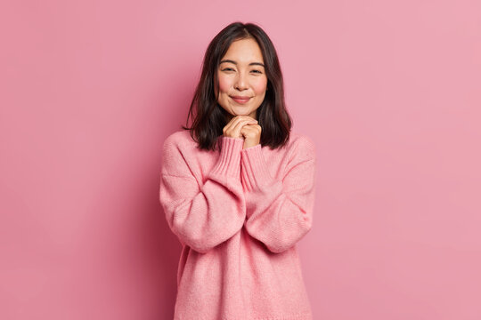 Charming brunette Asian girl with tender expression has rouge cheeks keeps hands under chin dressed in loose jumper poses against pink background. Attractive eastern woman has romantic feelings