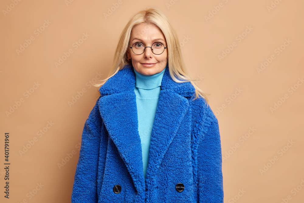 Wall mural Portrait of good looking middle aged woman looks calm at camera dresed in turtleneck blue winter coat round spectacles isolated over brown background. People outerwear seasonal clothes concept