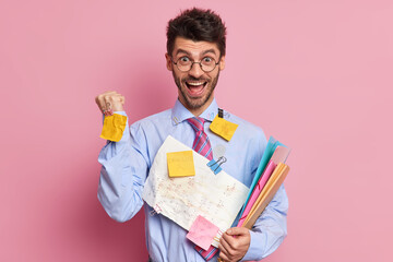 Successful businessman raises clenched fist and celebrates goals achievement poses with papers dressed in formal clothes isolated over pink background. Student finally finished working on report