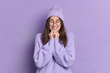 Pretty pleased brunette woman keeps hands under chin closes eyes and feels delighted glad to hear something good dressed in warm winter jumper hat big eyeglasses isolated over purple background