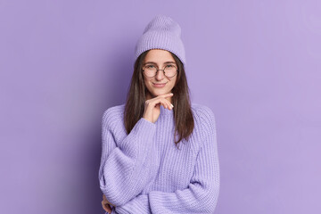 Brunette young beautiful woman holds chin and has sly expression tricky plan smiles pleasantly wears fashionable hat knitted sweater in one tone with background. Purple color monochrome shot