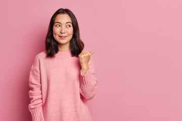 Charming pleased young Asian woman with rouge cheeks points aside on copy space shows advertisement...