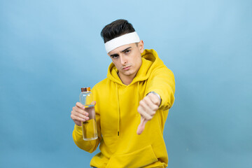 Young caucasian man wearing sportswear holding water bottle over isolated blue background with angry face, negative sign showing dislike with thumb down