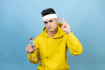 Young caucasian man wearing sportswear holding water bottle over isolated blue background gesturing finger crossed smiling with hope and looking side