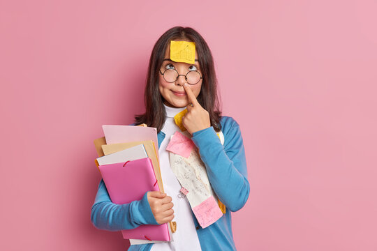 Funny nerdy student touches nose has sticky note with graphic stuck on forehead holds folders and papers concentrated above prepares for examination session. Woman studies papers with sums in office.