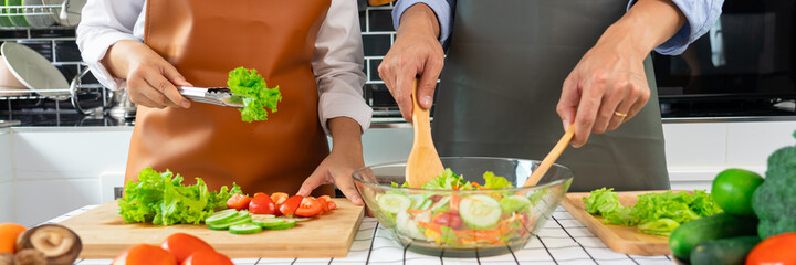Happy couple having fun standing in kitchen at home preparing vegetable salad husband and wife vegetarians chop vegetables prepare for dinner in loft kitchen at home.