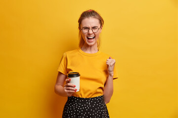 Emotional young European female shouts with widely opened mouth clenches fist drinks aromatic coffee drom disposable cup wears casual clothing and round spectacles isolated over yellow background