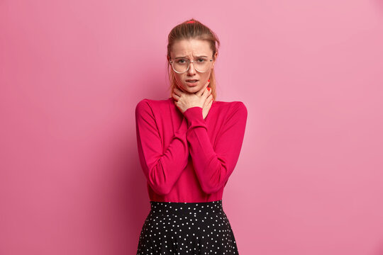 Unhappy girl suffocates because of painful strangle in throat keeps hands on neck cannot breath well has asthmatic attack health problems wears turtleneck skirt and spectacles isolated on pink wall