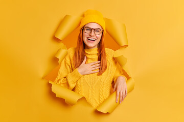 Positive ginger female model expresses sincere emotions keeps hand on chest feels gratitude smiles...