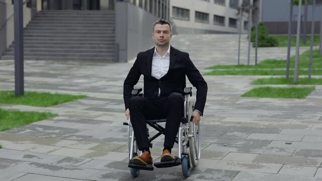 Portrait of serious man in formal suit looking to camera while sitting and going in wheel chair. Disabled successful businessman in 40s near office building. Concept of motivation.