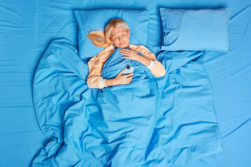 Photo of blonde senior woman uses mobile phone in bed fall asleep after watching video wears pajama sleeps peacefully. Middle aged female enjoys relaxation at bedroom. Bedding and napping concept