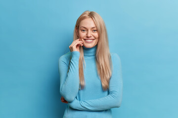 Fototapeta premium Monochrome shot of blonde pleased woman looks with gentle expression smiles happily wears turtleneck poses against blue background. Happy person has natural beauty. Emotions lifestyle concept