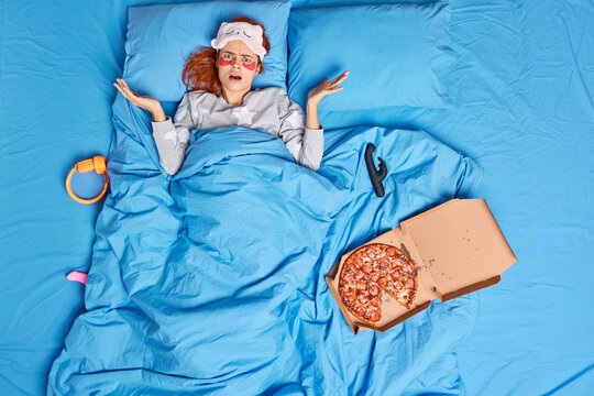 Indignant displeased woman shrugs shoulders and looks confused at camera lies in bed wears pajama sleepmask applies moisturizing patches under eyes surrounded with pizza sex toys headphones.